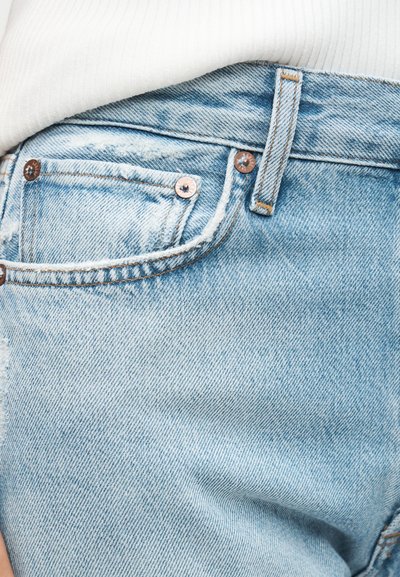 Κοντινή λήψη τσέπης από ανοιχτόχρωμο τζιν με χαλκομανίες και λευκό πουκάμισο περασμένο πάνω από τη μέση.