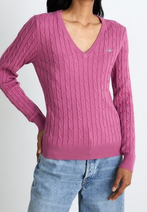 Personne portant un pull à col en V en maille torsadée rose avec des poignets et un ourlet côtelés, associé à un jean en denim bleu clair.