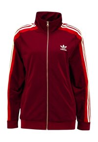 Röd träningsjacka med hög krage, fullt blixtlås och tre vita och röda ränder på varje ärm. Har Adidas-logotypen på bröstet.