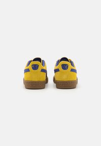 Gule mockasin-sneakers med blå detaljer, teksturert brun gummisåle og en liten logo på hælen; avrundet form og klassisk design.