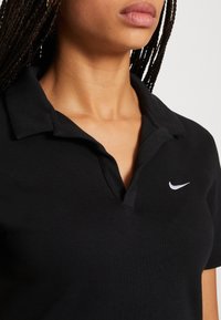 Črna polo majica iz bombaža s klasičnim ovratnikom, na levi strani prsnice pa je belo Nikeovo logo. Gladka tekstura, prilegajoča se zasnova.