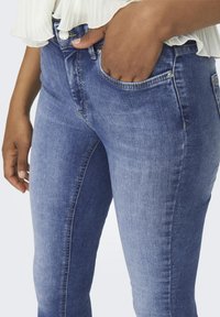 Jean en denim bleu avec une coupe slim et des coutures visibles. Il possède une poche frontale, une fermeture à bouton et une texture douce.
