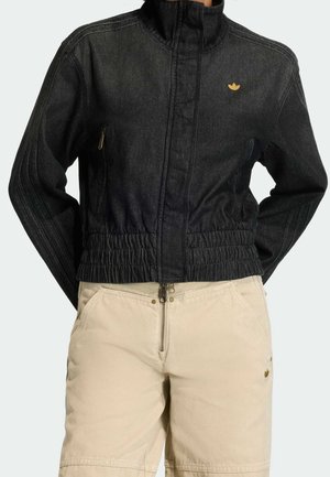 Person trägt eine schwarze Jacke mit Reißverschluss und elastischem Bund sowie beige hoch taillierte Hosen mit Frontreißverschluss und Taschen.
