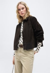 Νεαρή γυναίκα με μαύρο σακάκι με κουμπιά πάνω από λευκό πουκάμισο μακρυμάνικο με μαύρες πουά βούλες, μπεζ παντελόνι και καφέ ζώνη.