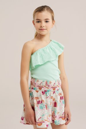 Jeune fille portant un haut asymétrique vert menthe à volants et une jupe colorée à motifs, debout sur un fond neutre.