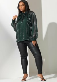Blusa verde oscuro y brillante con un lazo en el cuello; mangas abullonadas transparentes. Pantalones de cuero ajustados negros y sandalias negras con tiras.