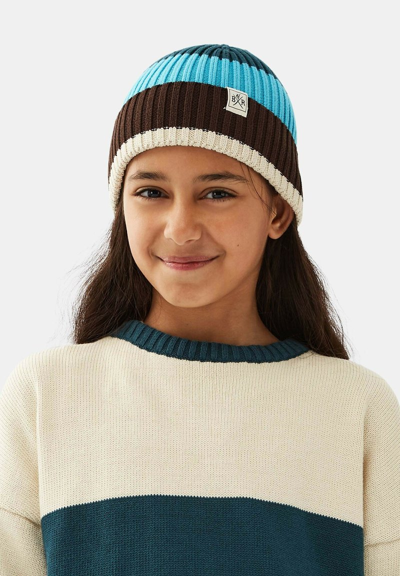 Strikket hue i blå, brun og creme striber. Har ribbet tekstur og et logo-patch. Kombination med en sweater i creme og teal.