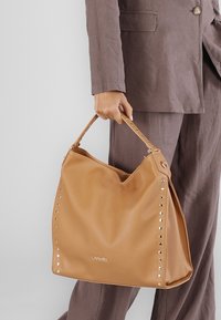 Brun läder hobo-väska med en slapp design, prydd med silvernitar längs ena sidan och en enkel axelrem.
