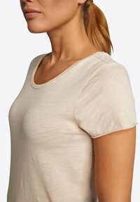 Helles beige T-Shirt mit kurzen Ärmeln aus weichem Stoff, mit rundem Halsausschnitt und subtilen Details an den Ärmeln. Glatte Textur.