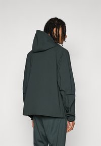 Hooded jacket in dark green, made from lightweight material, with a loose fit and elastic cuffs, featuring minimal detailing and a plain design.