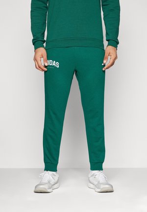 Mand iført grønne Adidas træningsbukser og matchende sweatshirt med hvide sneakers, stående mod en ensfarvet baggrund.