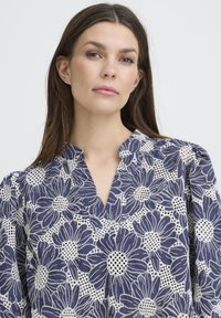 Blouse bleu marine à imprimé floral blanc, avec un décolleté fendu et des manches bouffantes. Le tissu présente un aspect texturé et transparent avec un motif détaillé.