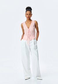 Rosa ärmelloses Top mit V-Ausschnitt und Frontbindedetail, kombiniert mit locker sitzenden weißen Hosen und spitzen weißen Schuhen. Sauberes Design.