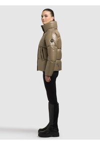 Khaki pikowana kurtka z wysokim kołnierzem, błyszczącą fakturą, boczną kieszenią i naszywką z logo. Noszona z czarnymi dopasowanymi spodniami i wysokimi kozakami do kolan.