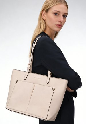 Donna bionda con blazer nero che porta una grande borsa tote beige in pelle con due tasche frontali e tracolle regolabili.
