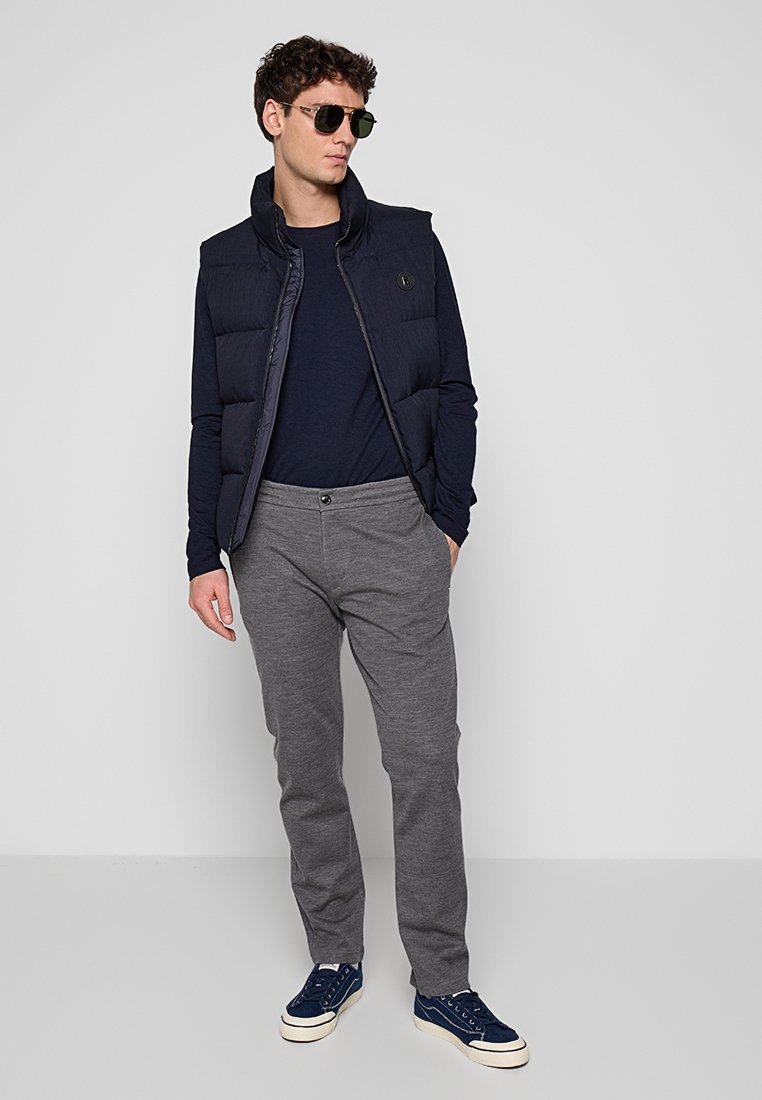 Marineblauer, gepolsterter Weste über einem marineblauen Langarmshirt, grauen Hosen und blauen Sneakers. Accessoires sind dunkel getönte Sonnenbrillen.