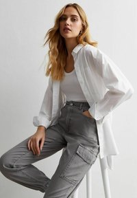 Ung kvinna med långt blont hår, klädd i en vit skjorta över ett vitt linne och grå cargobyxor, sitter på en vit pall mot en enfärgad bakgrund.