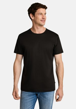 Mann trägt schlichtes schwarzes Kurzarm-T-Shirt und blaue Jeans, steht und schaut seitlich vor einem einfachen hellen Hintergrund.