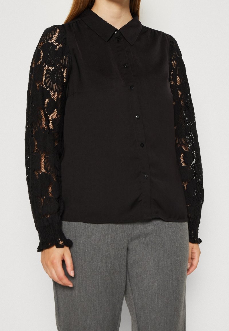 Femme portant un chemisier noir boutonné avec des manches longues en dentelle florale et un pantalon gris, debout devant un fond blanc.