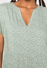 Eine Frau trägt ein hellgrünes Kleid mit kleinen weißen Blattmustern und einem V-Ausschnitt; ihr geflochtenes Haar ist teilweise sichtbar.