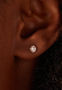 Boucles d'oreilles en zircon cubique transparent, rondes, montées sur un support en métal doré, fixées à une oreille brun foncé, mettant en valeur une texture lisse et un éclat.
