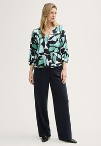 Blusa multicolor con un patrón abstracto de hojas en negro, verde y blanco, combinada con pantalones de pierna ancha en azul marino y zapatos de punta negra.