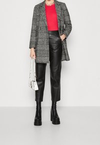 Frau in einem schwarz-weißen karierten Mantel, rotem Oberteil, schwarzen Lederhosen, Plateauboots und mit einer weißen Handtasche, die vor einem einfarbigen Hintergrund steht.