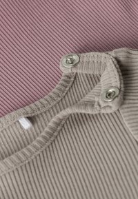 Lähivõte soonilisest kangast, millel on trukid, näidates trukikinnituse detaili beežikal riietusel tuhmi roosa tausta ees.