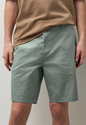 Homme portant un short vert clair jusqu'aux genoux et un t-shirt marron, debout les mains relâchées de chaque côté.