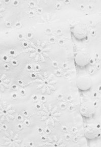 Vitt broderat tyg med blommönster, med små knappproppar för stängning. Strukturerad yta med öppna vävningar och intrikata detaljer.