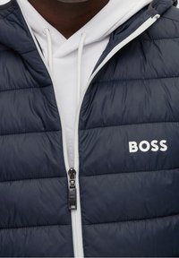 Detalní záběr na osobu v námořnické prošívané bundě s bílým zipem a logem BOSS přes bílou mikinu s tkaničkami.