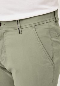 Hellgrüne Baumwollhosen mit einer Fronttasche, subtilen Stichdetails und einem Reißverschluss mit einem markierten Ziehteil.