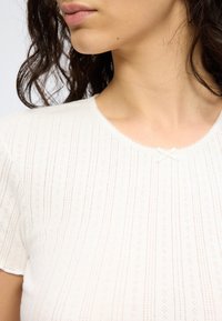 Κοντινό πλάνο στον λαιμό και τον ώμο μιας γυναίκας που φοράει ένα λευκό πουκάμισο με υφή και ένα μικρό διακοσμητικό φιόγκο στο ντεκολτέ.