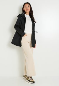 Mujer vestida con una chaqueta negra con capucha, una blusa blanca, pantalones anchos beige y zapatillas con estampado, de pie contra una pared blanca.