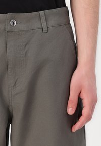 Grijze, lichte broek met een gladde textuur, voorzien van een standaard tailleband en een metalen knoopsluiting. Stiksels zichtbaar langs de naden.