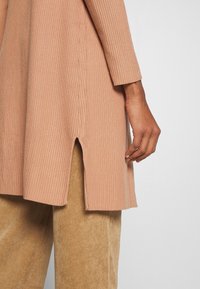 Rippenshirt mit langen Ärmeln in sanftem Pfirsichton, mit einem seitlichen Schlitze und kontrastierenden beigen Cordhosen. Strukturierter, taillierter Schnitt mit klassischer Silhouette.