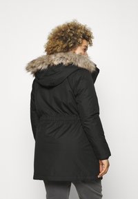 Parka noire avec une capuche doublée de fourrure, taille cintrée et manches longues. Il présente une texture lisse et une longueur jusqu'au mi-cuisse.