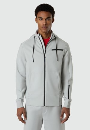 Uomo che indossa una felpa con cappuccio grigio chiaro con zip e cordini neri, con una maglietta rossa sotto, in piedi davanti a uno sfondo semplice.