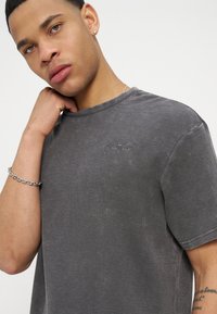 Νεαρός με κοντά μαλλιά που φοράει γκρι σκουρόχρωμο T-shirt με υφή, ασημένιο βραχιόλι αλυσίδα, μικρό σκουλαρίκι και αγγίζει τον λαιμό του με ουδέτερη έκφραση.