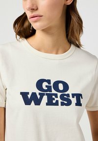Camiseta de algodón color crema con mangas cortas que presenta un texto en negrita de color azul marino que dice "GO WEST". Cuello redondo clásico.