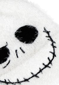 Close-up van een wit fuzzy stof met een zwart geborduurd gezicht dat op een schedel lijkt, met holle ogen en een geborduurde glimlach.