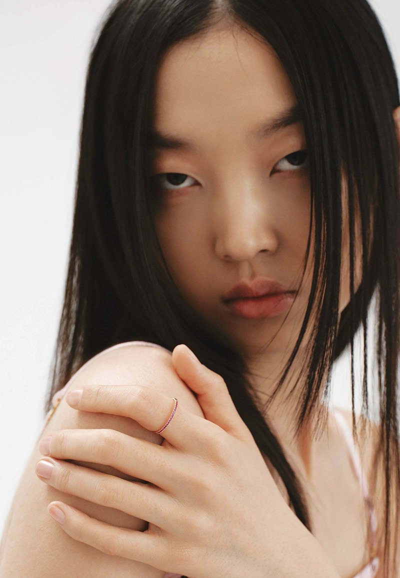 Primer plano de una mano con un anillo de banda delgada y colorido. El fondo es neutro y el sujeto tiene el cabello largo y oscuro, con una textura de piel suave.