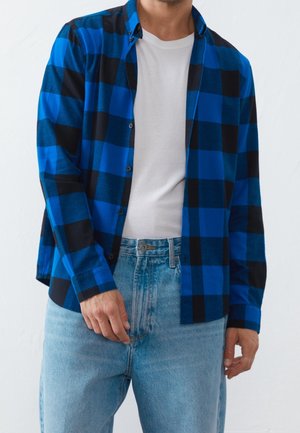 Mężczyzna w niebiesko-czarnej koszuli w kratę na białym t-shircie i jasnoniebieskich dżinsach, stojący na tle prostego jasnego tła.
