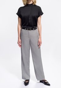 Femme portant un blouse noire à manches courtes, un pantalon large gris avec une ceinture noire, et des ballerines noires à bouts pointus avec des lanières.