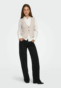 Gilet en laine gris clair avec deux boutons sur le devant, porté sur une chemise blanche à col, associé à un jean noir taille haute et des chaussures noires.