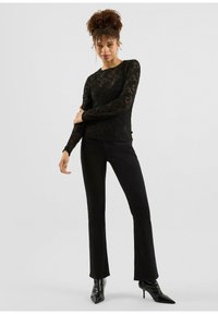 Schwarzes Spitzenoberteil mit langen Ärmeln und Blumendessin, kombiniert mit schwarzen Schlaghosen und spitzen schwarzen Stiefeln. Elegante Silhouette und strukturierte Details.