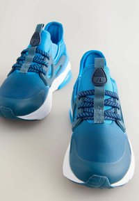 Los zapatos deportivos azules presentan una parte superior textil lisa con paneles de malla, cordones elásticos y un cuello acolchado. La suela con dibujo proporciona tracción.