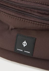 Ruskea kangaskassi, jossa useita päävetoketjuja, teksturoitu pinta ja musta etiketti, jossa lukee "SAMSØE SAMSØE" sekä ympyräembleemi.