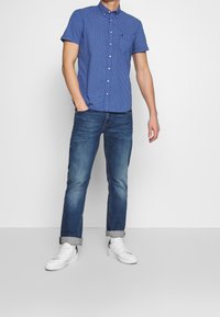 Camicia azzurra a maniche corte con piccoli pois bianchi, colletto button-down, taschino sul petto, abbinata a jeans blu e sneaker bianche.
