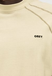Κοντινό πλάνο μπεζ μπλούζας με στρογγυλή λαιμόκοψη, με ορατές καφέ ραφές και μικρό μαύρο λογότυπο "OBEY" στο στήθος.